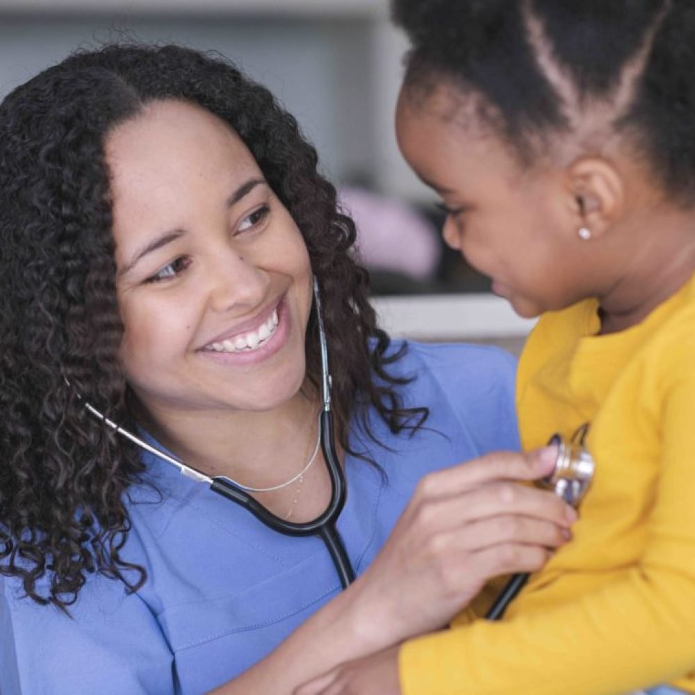 A female doctor of African descent is doing a routine check-up on a toddler. The medical professional is using a stethoscope to check the little girl’s heart rate. The child is seated on a medical exam table. The doctor is smiling warmly at her patient.